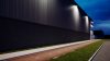 Un potente proyector LED exterior montado en el lateral de un edificio, proyectando una luz blanca brillante sobre un área de estacionamiento al atardecer.